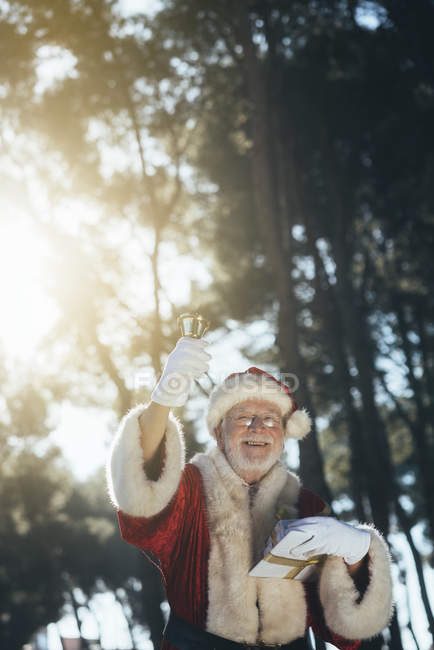 Cheerful senior man in costume of Santa Claus standing with present and bell in gloved hands on nature background — Stock Photo