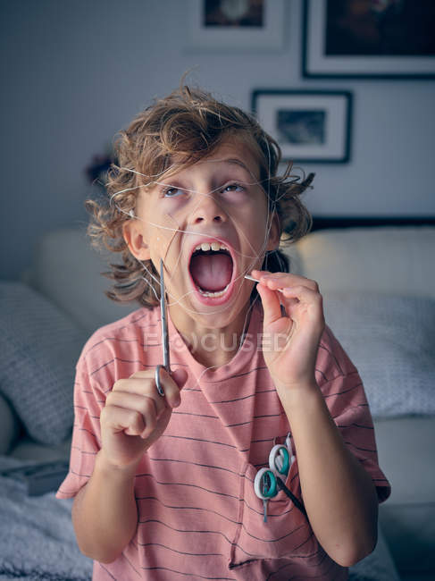 Excited curly kid pulling dental floss for which milk tooth tied with scissors in hand at home looking up — Stock Photo