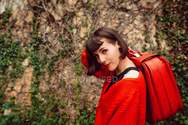 Donna in rosso con grande valigia rossa posa contro muro di pietra con piante — Foto stock