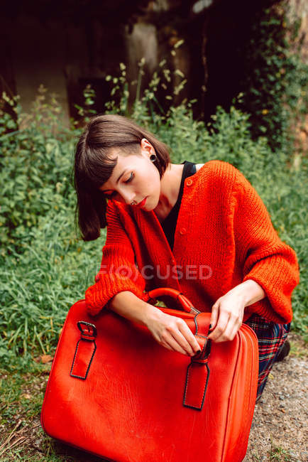 Jeune femme avec ceinture de contrôle de cheveux élégant sur valise rouge assis dans la nature — Photo de stock