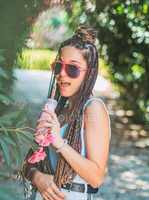 Весела молода жінка в сонцезахисних окулярах з дредлоками п'є коктейль і дивиться на камеру в парку — стокове фото