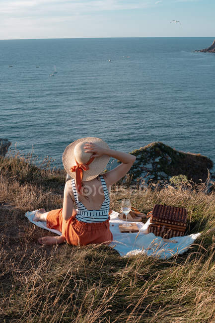 Vue arrière de la femme en chapeau avec boisson assise sur le rivage et regardant romantiquement le long de l'eau sereine — Photo de stock
