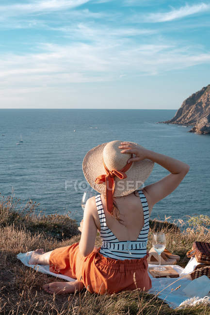 Visão traseira da mulher de chapéu com bebida sentada na costa e romanticamente olhando ao longo da água serena — Fotografia de Stock