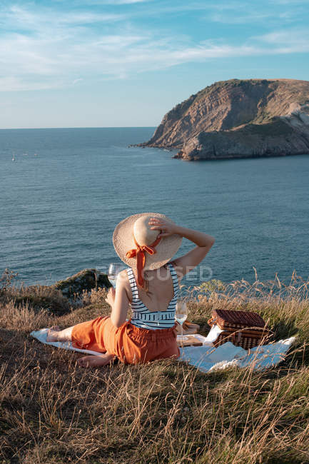 Visão traseira da mulher de chapéu com bebida sentada na costa e romanticamente olhando ao longo da água serena — Fotografia de Stock