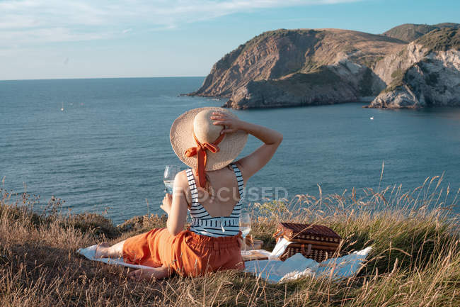 Rear view of woman in hat with drink sitting on shore and romantically looking along to serene water — Stock Photo