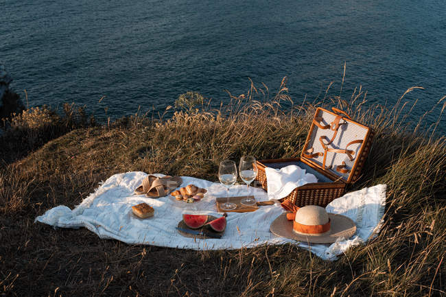 Коврик для пикника с романтическим набором с бокалами напитков и едой на сухом берегу с безмятежной водой — стоковое фото