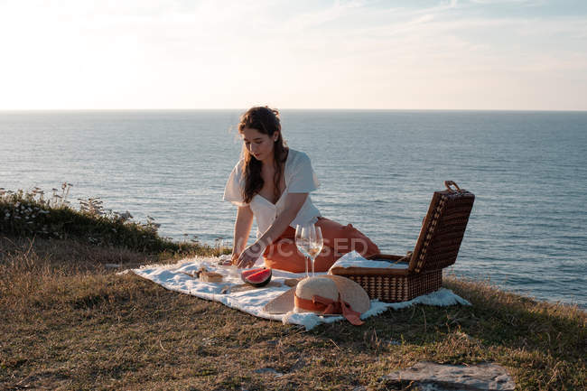 Side view of woman sitting on mat for picnic eating on dry shore and serene water and hills — Stock Photo