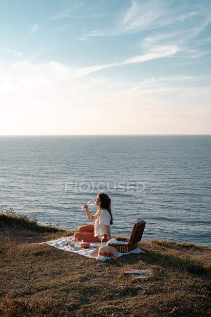 Side view of woman with drink sitting on mat for picnic on dry shore and romantically with eyes closed near serene water and hills — Stock Photo