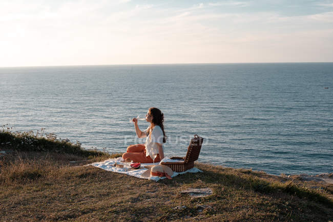 Vista lateral da mulher com bebida sentada no tapete para piquenique na costa seca e romanticamente com os olhos fechados perto de água serena e colinas — Fotografia de Stock