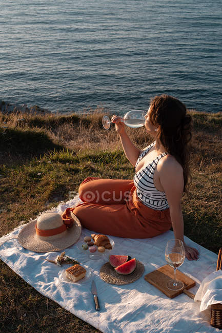 Joven mujer románticamente beber vino en la orilla cerca de sereno agua y colinas - foto de stock