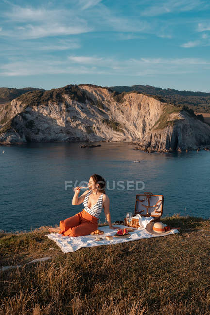 Молодая женщина романтически пьет вино на берегу у безмятежной воды и холмов — стоковое фото