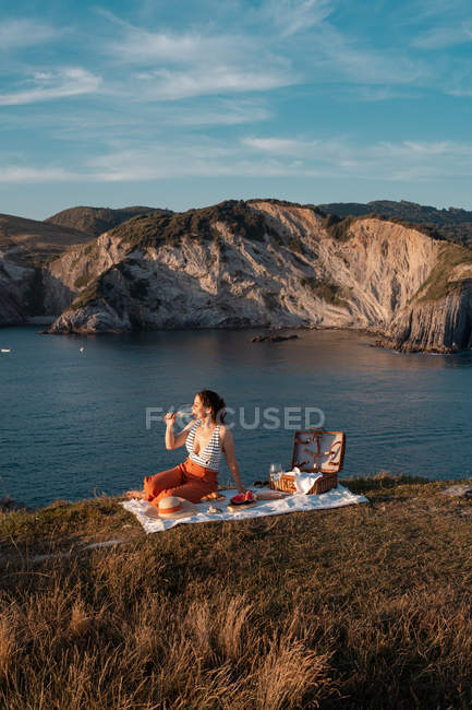 Молода жінка романтично п'є вино на березі біля спокійної води і пагорбів — стокове фото