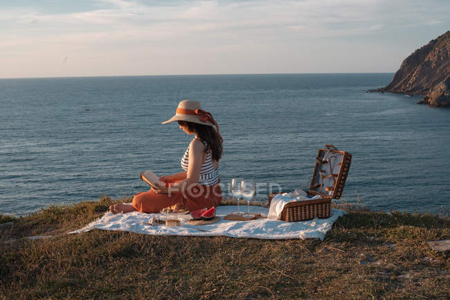 Side view of woman in hat with book sitting on mat for picnic with watermelon — Stock Photo