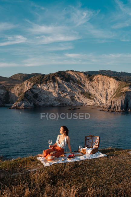 Seitenansicht einer Frau, die sich bei einem Drink auf einer Matte entspannt, um am ruhigen Wasser und in den Hügeln zu picknicken — Stockfoto
