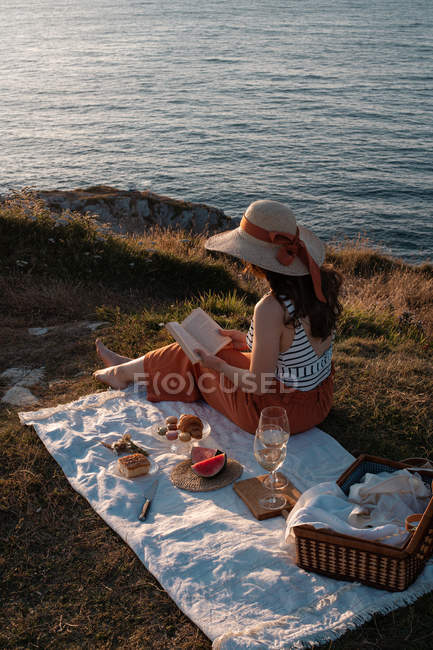 Femme dans le livre de lecture de chapeau sur le tapis pour pique-nique sur la rive sèche par l'eau sereine — Photo de stock