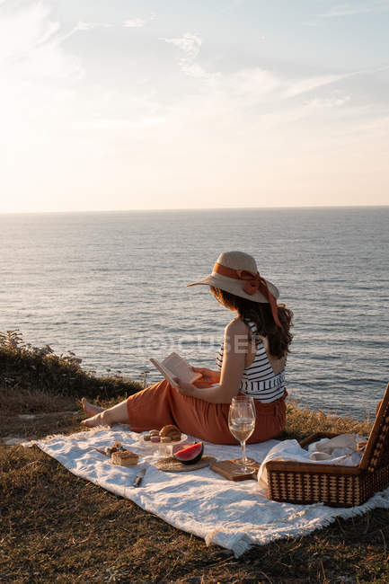 Mulher em chapéu livro de leitura no tapete para piquenique na costa seca por água serena — Fotografia de Stock