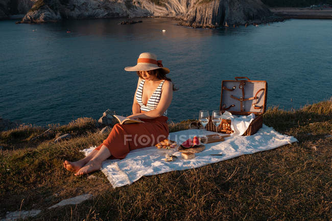 Femme en chapeau lecture livre assis sur le tapis pour pique-nique dans la lumière du soleil — Photo de stock