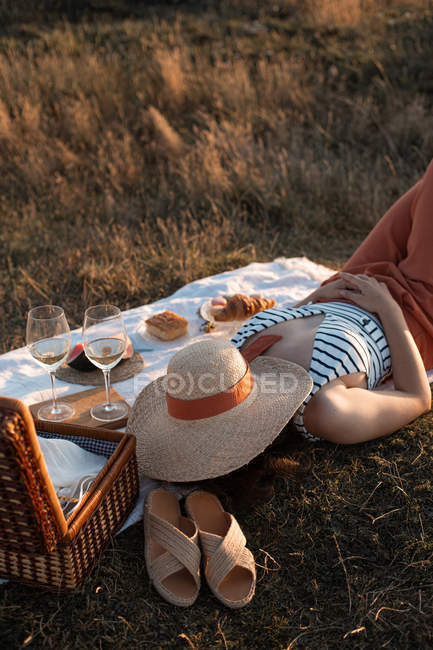 Dall'alto donna che si diverte sdraiata su un tappeto da picnic bianco con cappello sul cestino vicino sul prato — Foto stock