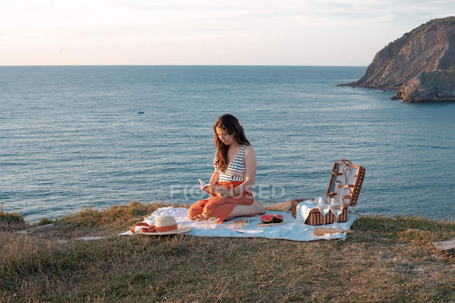 Side view of woman with book sitting on mat for picnic with watermelon on dry shore — Stock Photo