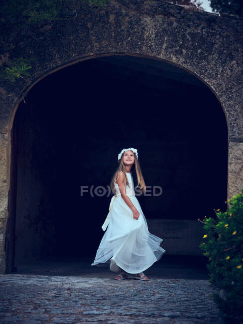 Очаровательная маленькая девочка в белом платье и цветочном браслете танцует под аркой в парке, глядя вверх — стоковое фото