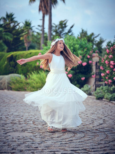 Cute little girl in white airy dress and flower headband spinning in park — Stock Photo