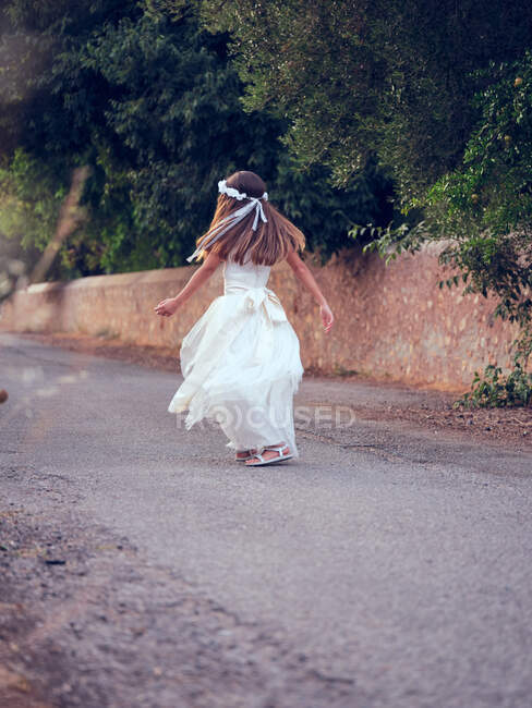 Vue arrière de la petite fille mignonne méconnaissable en robe blanche aérée et bandeau de fleurs marchant dans le parc — Photo de stock