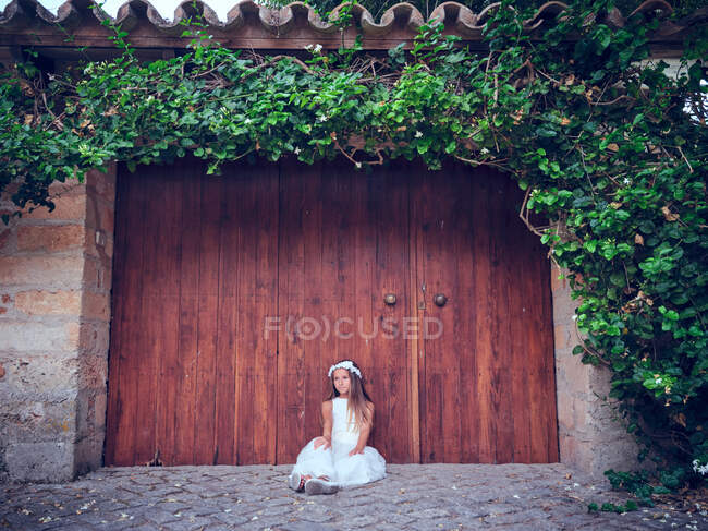 Cute little girl in white long dress and headband resting sitting near wooden door with ivy looking away — Stock Photo