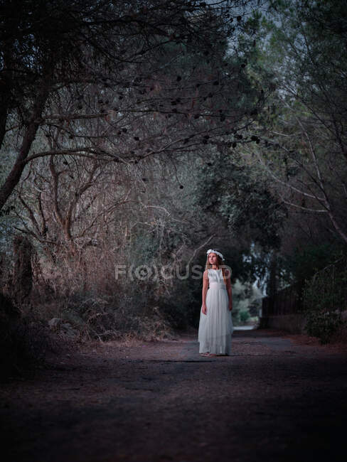 Маленькая одинокая девочка в длинном белом платье стоит на дороге в темном переулке и смотрит в сторону — стоковое фото