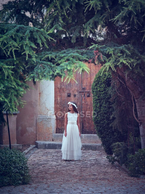 Adorable little girl in airy long white dress and flower headband walking under arch in park looking away — Stock Photo