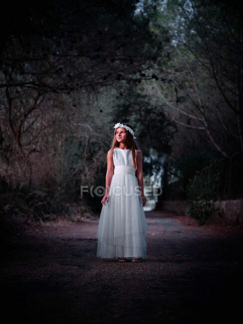 Pequena menina solitária em vestido branco longo em pé na estrada no beco escuro olhando para longe — Fotografia de Stock
