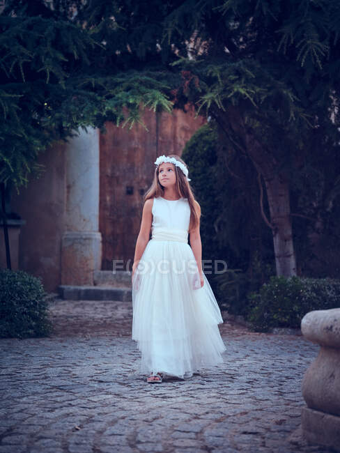 Чарівна маленька дівчинка в повітряній довгій білій сукні та квітковій пов'язці, що йде під аркою в парку, дивлячись подалі — стокове фото