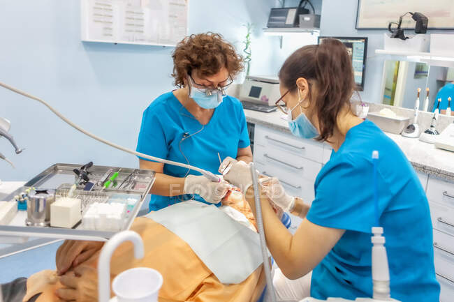 Especialistas seguros en guantes quirúrgicos poniendo sello y trabajando con herramientas dentales en boca del paciente acostado en silla en gabinete - foto de stock