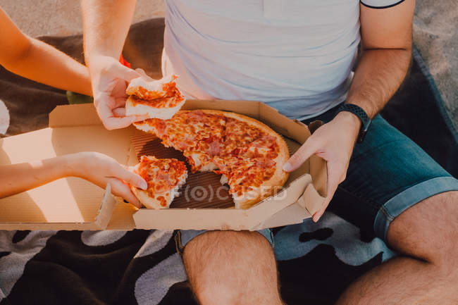 Обрізаний вид на невпізнавану пару, що сидить на пляжі для рушників на піску та їсть піцу — стокове фото