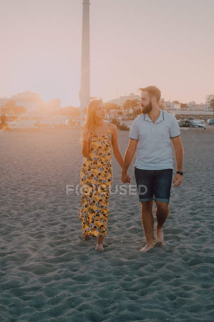 Giovani felici innamorati che camminano guardando l'un l'altro e tenendosi per mano scalzi in riva al mare alla luce del sole — Foto stock