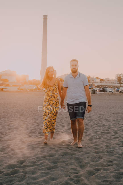 Giovani felici innamorati che camminano guardando l'un l'altro e tenendosi per mano scalzi in riva al mare alla luce del sole — Foto stock