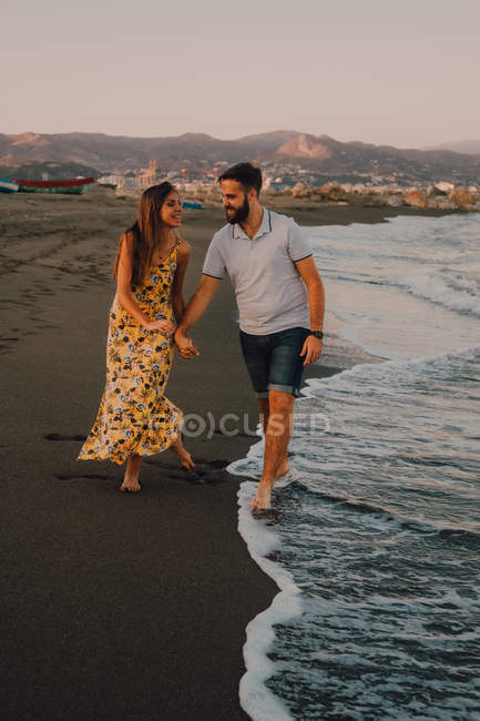 Jóvenes felices enamorados caminando mirándose unos a otros y tomándose de las manos descalzos en la playa a la luz del sol - foto de stock