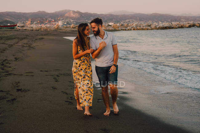 Giovani felici innamorati che camminano guardando l'un l'altro e tenendosi per mano scalzi in riva al mare alla luce del sole — Foto stock