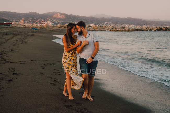 Happy young people in love walking looking to each other and holding hands barefoot in seaside in sunlight — Stock Photo