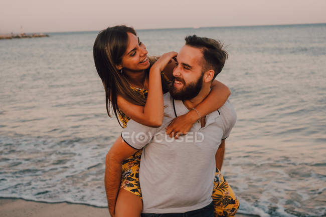 Bearded man giving woman piggyback ride in foamy water seaside — Stock Photo