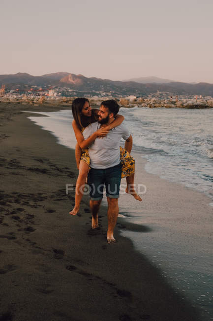 Barbudo hombre dando mujer a cuestas paseo en agua espumosa playa - foto de stock