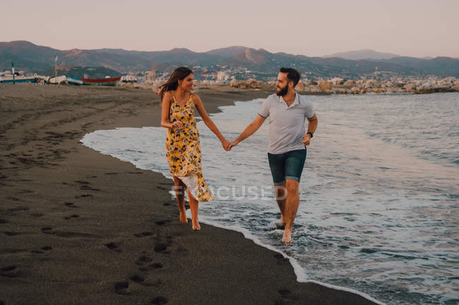 Heureux les jeunes amoureux marchant en regardant les uns vers les autres et se tenant les mains pieds nus au bord de la mer au soleil — Photo de stock