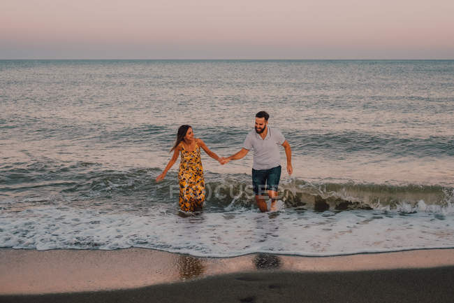 Happy young people in love running looking to each other and holding hands barefoot in seaside in sunlight — Stock Photo