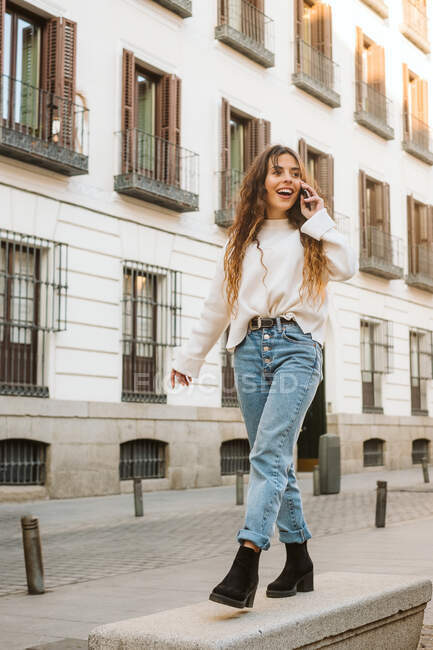 Young woman in stylish outfit smiling and answering phone call while walking on border on city street — Stock Photo