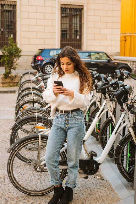 Fröhliche junge Frau in lässigem Outfit sitzt auf Leihfahrrädern an der Sammelstelle und blickt in die Kamera auf der Straße — Stockfoto