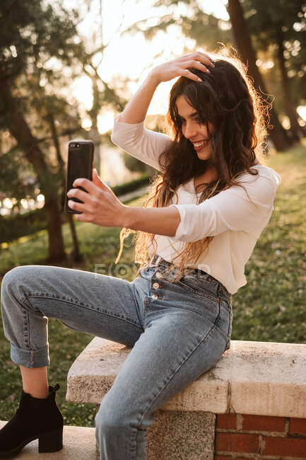 Jovem senhora em roupa casual sorrindo e tocando o cabelo enquanto sentado na fronteira no parque e tomando selfie no dia ensolarado — Fotografia de Stock