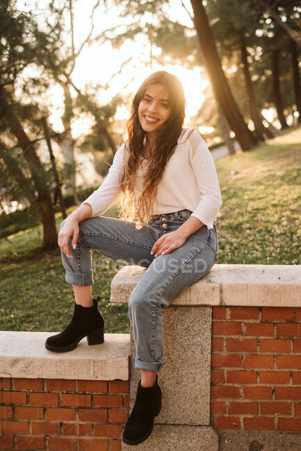 Pretty young female in casual outfit smiling and looking at camera while leaning on border on sunny day in park — Stock Photo