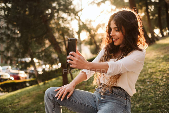 Giovane signora in abito casual sorridente e toccante capelli mentre seduto sul confine nel parco e prendere selfie nella giornata di sole — Foto stock