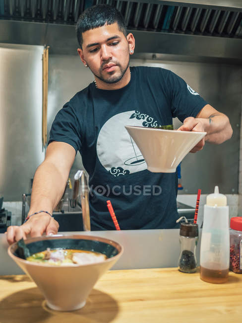 Junger Mann in schwarzem T-Shirt legt auf den Tisch Schüsseln mit frisch gekochtem japanischem Gericht namens Ramen — Stockfoto