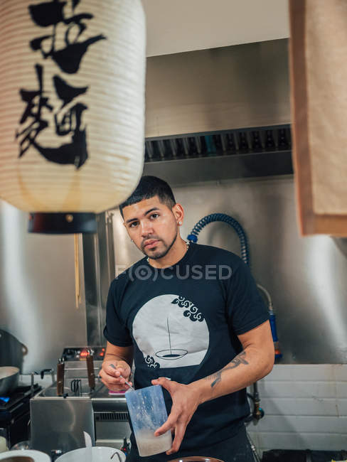 Joven cocina masculina étnica plato japonés llamado ramen y mirando en la cámara en la cafetería asiática - foto de stock