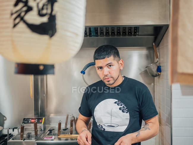 Молодой этнический мужчина готовит японское блюдо и смотрит в камеру в азиатском кафе — стоковое фото